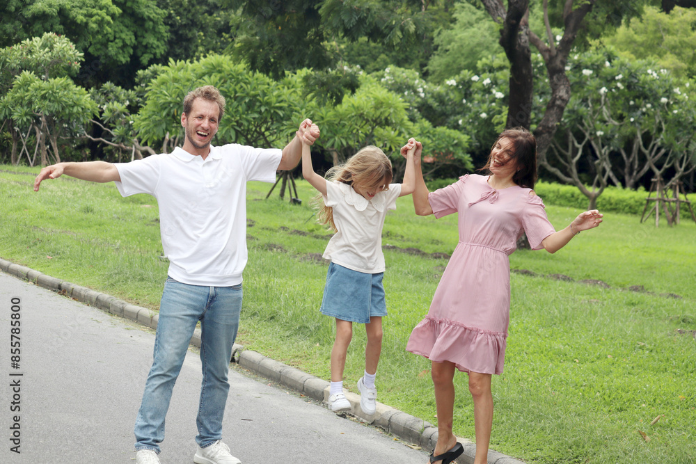 Fototapeta premium Father, mother and cute kid daughter taking a walk together in beautiful summer garden. Joyful parents hold hands, carry and lift up girl child on the park walkway at green park. Happy family outside.