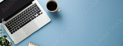 An office desk with a laptop, notebooks, and coffee cup on a blue color background. A panoramic banner background with space for a copy.