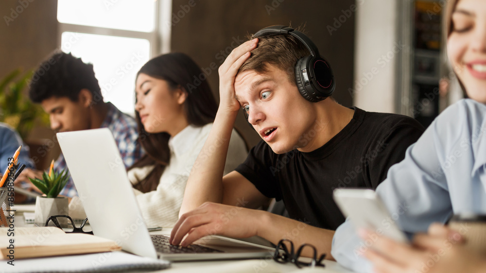 Shocked student guy looking test results on laptop, studying with classmates in library