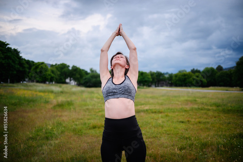 Woman practicing yoga in a wide-open field, reaching her arms upward towards the cloudy sky. Green trees and hills are visible in the background.