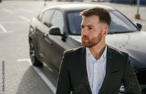Focused portrait, against vehicle. Businessman in suit is near his black car outdoors © standret