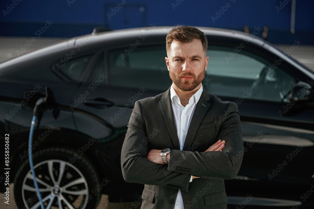 Front view, arms crossed. Businessman in suit is near his black car outdoors Front view, arms crossed. Businessman in suit is near his black car outdoors