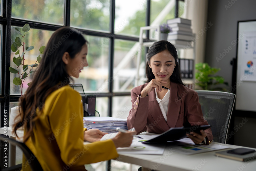 Two women are sitting at a desk, one of them is looking at a tablet