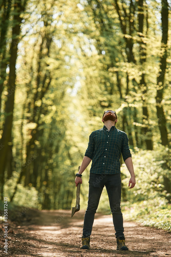 Looks up, holding an axe. Bearded man is in the forest at daytime Looks up, holding an axe. Bearded man is in the forest at daytime