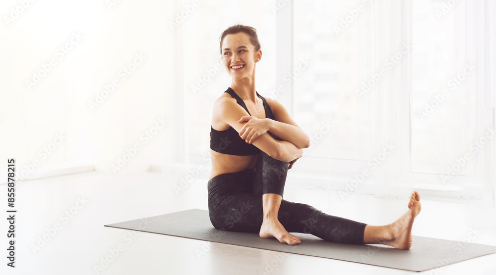 A young woman is seated on a yoga mat performing a stretch in a brightly sunlit room. She appears relaxed and focused during her morning exercise routine. A young woman is seated on a yoga mat performing a stretch in a brightly sunlit room. She appears relaxed and focused during her morning exercise routine.