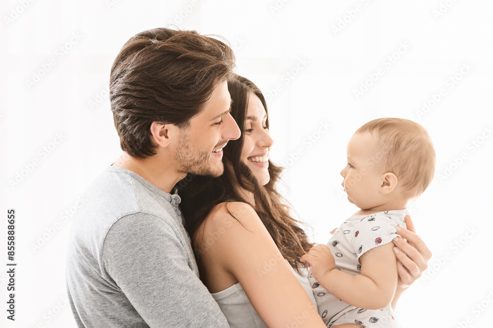 A happy family of three is pictured against a white background. The mother is holding the baby boy, who is looking at his parents. They are all smiling at the baby, showing love and affection. A happy family of three is pictured against a white background. The mother is holding the baby boy, who is looking at his parents. They are all smiling at the baby, showing love and affection.