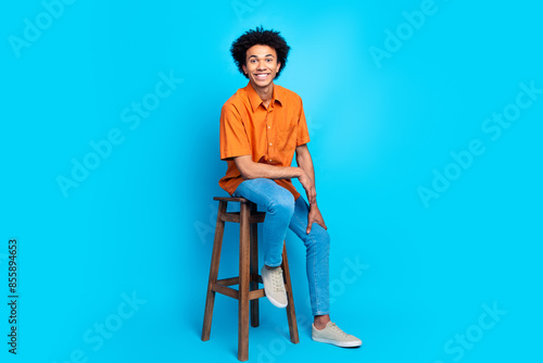 Full length photo of attractive funny guy wear orange shirt sitting chair emtpy space isolated blue color background