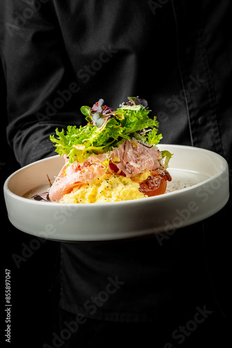 Fototapeta Naklejka Na Ścianę i Meble -  A close-up of a gourmet dish featuring scrambled eggs and smoked salmon, garnished with fresh herbs, presented in a white bowl held by a person in black attire against a dark background