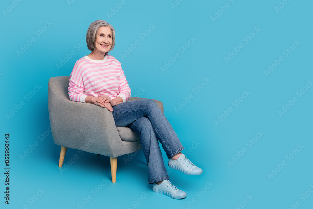 Full size photo of attractive retired woman sit armchair look empty space dressed stylish striped clothes isolated on blue color background Full size photo of attractive retired woman sit armchair look empty space dressed stylish striped clothes isolated on blue color background