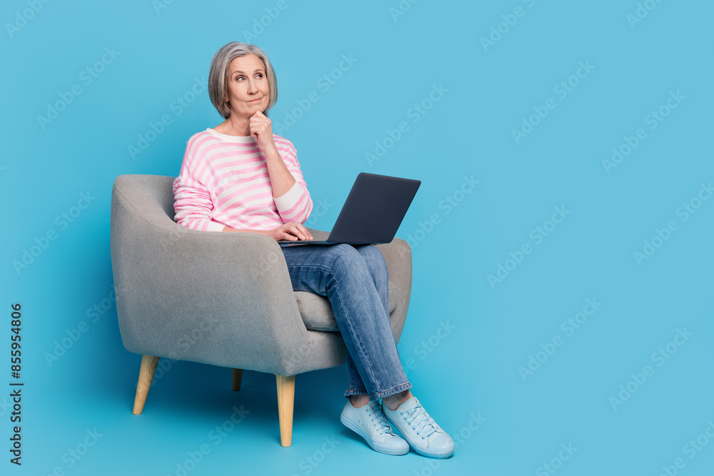 Full size photo of retired woman sit armchair netbook look minded empty space dressed striped clothes isolated on blue color background Full size photo of retired woman sit armchair netbook look minded empty space dressed striped clothes isolated on blue color background