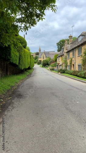 Vertical shot. Personal POV walking in village street with yellow stone houses on one side and tall hedgerow on other in Cotswolds, England.