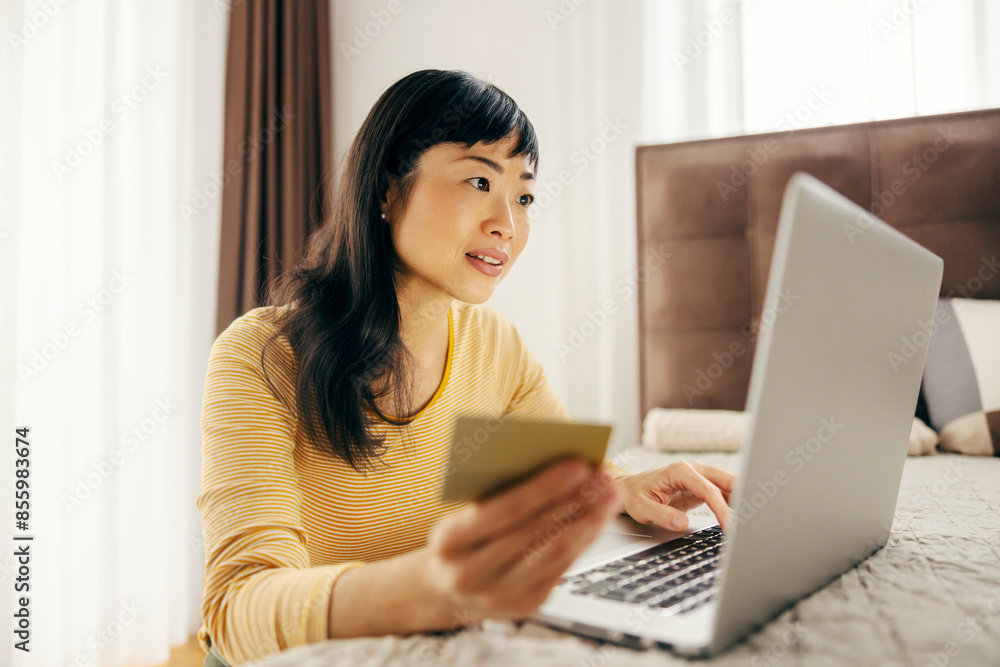 An asian woman using credit card and a laptop for online shopping in a bedroom. An asian woman using credit card and a laptop for online shopping in a bedroom.
