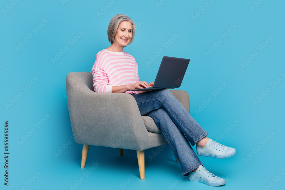 Full length photo of lovely pensioner lady sit armchair netbook dressed stylish striped garment isolated on blue color background Full length photo of lovely pensioner lady sit armchair netbook dressed stylish striped garment isolated on blue color background