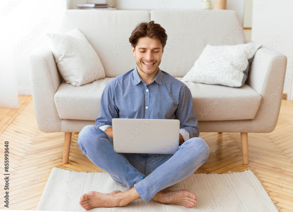 A man is sitting on the floor in a living room, using a laptop. He is smiling and appears to be relaxed. A man is sitting on the floor in a living room, using a laptop. He is smiling and appears to be relaxed.