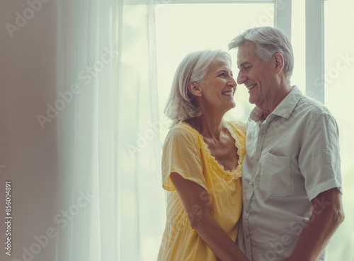 Coppia felice di uomini anziani abbracciati vicini alla finestra, uomo e donna, che si amano e sorridono. Vicini, lei con abito giallo e lui camicia bianca. photo