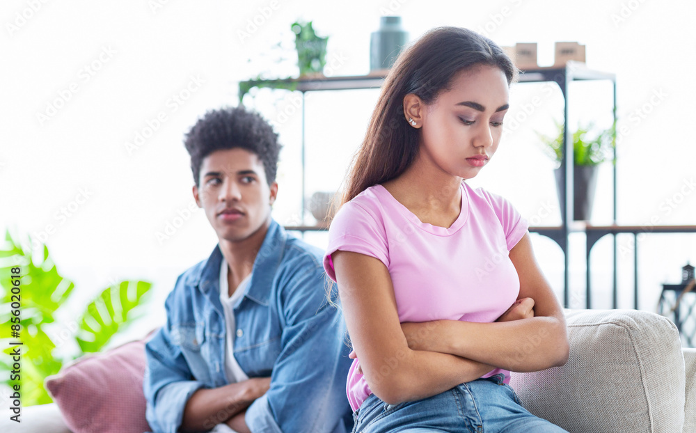 Black teen couple sits on a couch in a living room. They are both looking away from each other and appear to be upset. The girl has her arms crossed and is looking down Black teen couple sits on a couch in a living room. They are both looking away from each other and appear to be upset. The girl has her arms crossed and is looking down