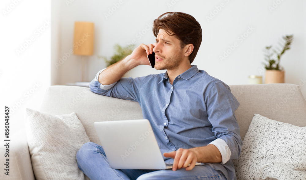 A man sits on a couch, talking on a phone while working on a laptop.
