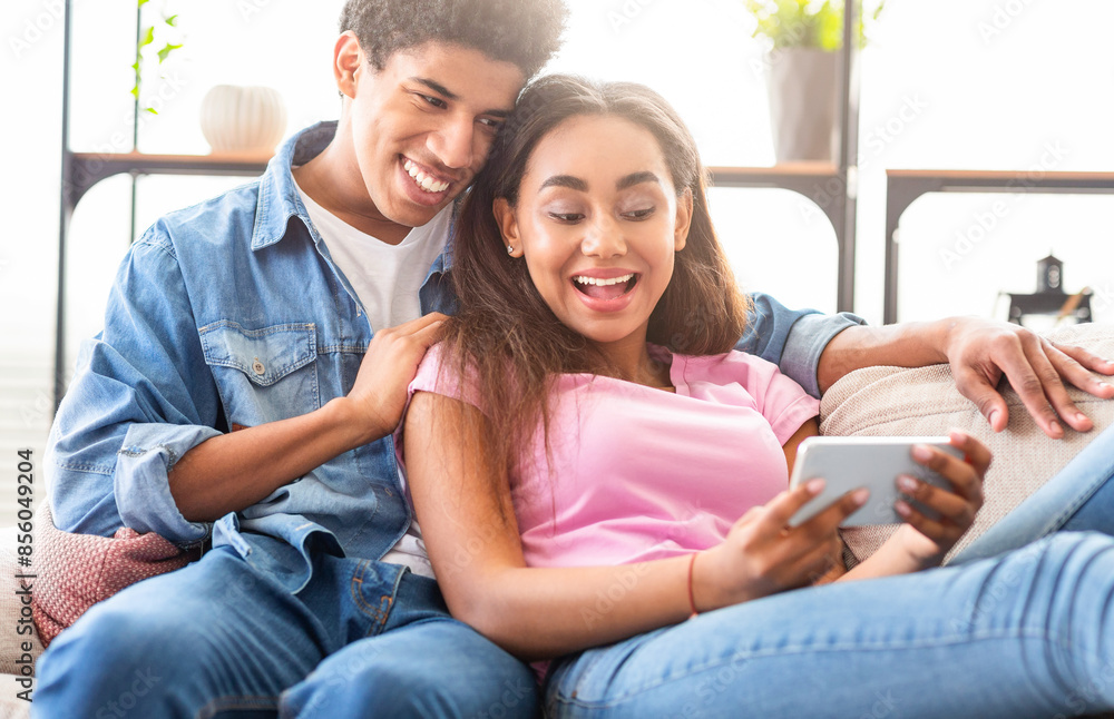 African American teen guy and girl are sitting on a couch, the girl is looking at her smartphone while the guy looks at her with a smile. Both are relaxed and comfortable, closeup African American teen guy and girl are sitting on a couch, the girl is looking at her smartphone while the guy looks at her with a smile. Both are relaxed and comfortable, closeup