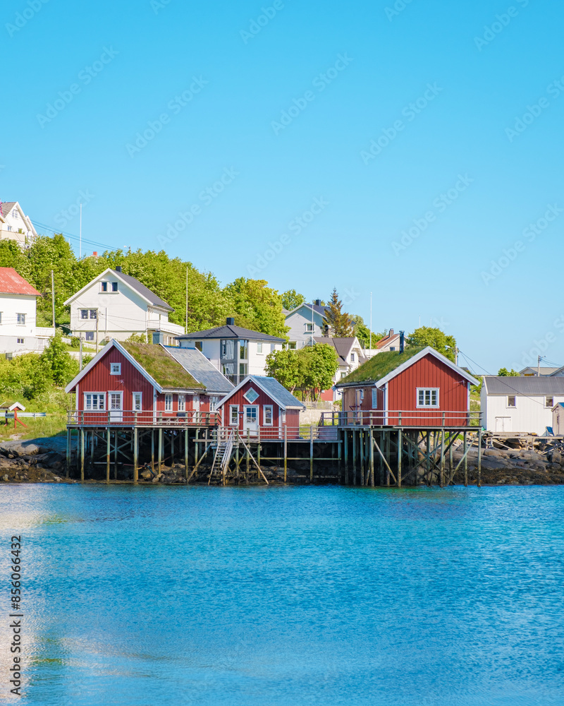 A scenic view of red cabins nestled in a picturesque Norwegian town. The houses are perched on the edge of the water, with a stunning blue sky and calm water in the background. Reine, Lofoten, Norway A scenic view of red cabins nestled in a picturesque Norwegian town. The houses are perched on the edge of the water, with a stunning blue sky and calm water in the background. Reine, Lofoten, Norway
