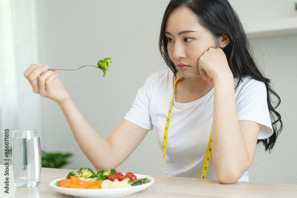 unhappy asian women is on dieting time looking at broccoli on the fork. girl do not want to eat vegetables and dislike taste of broccoli. unhappy asian women is on dieting time looking at broccoli on the fork. girl do not want to eat vegetables and dislike taste of broccoli.