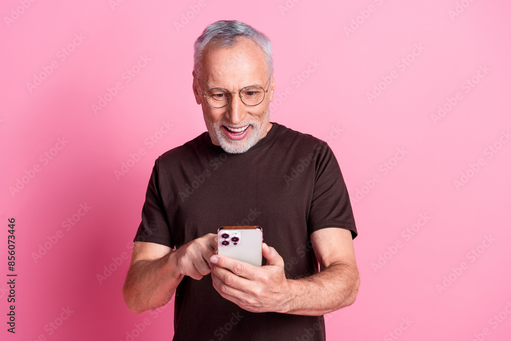 Portrait of positive man with beard dressed t-shirt in glasses typing message look at smartphone isolated on pink color background Portrait of positive man with beard dressed t-shirt in glasses typing message look at smartphone isolated on pink color background