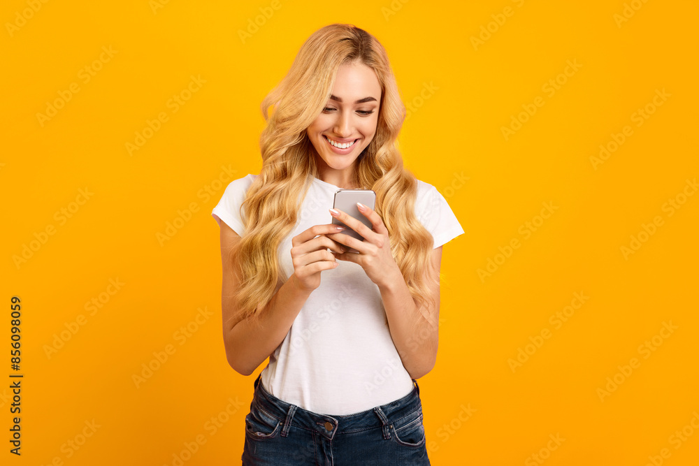 A young woman with long blonde hair, wearing a white t-shirt and blue jeans, is smiling and looking at her smartphone in front of a bright yellow background. She appears happy and engaged A young woman with long blonde hair, wearing a white t-shirt and blue jeans, is smiling and looking at her smartphone in front of a bright yellow background. She appears happy and engaged