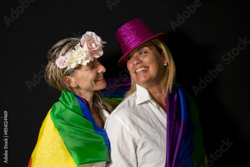 Two blonde women wearing the gay pride flag, a flower crown and a pink top hat look at each other knowingly on a black background. photo