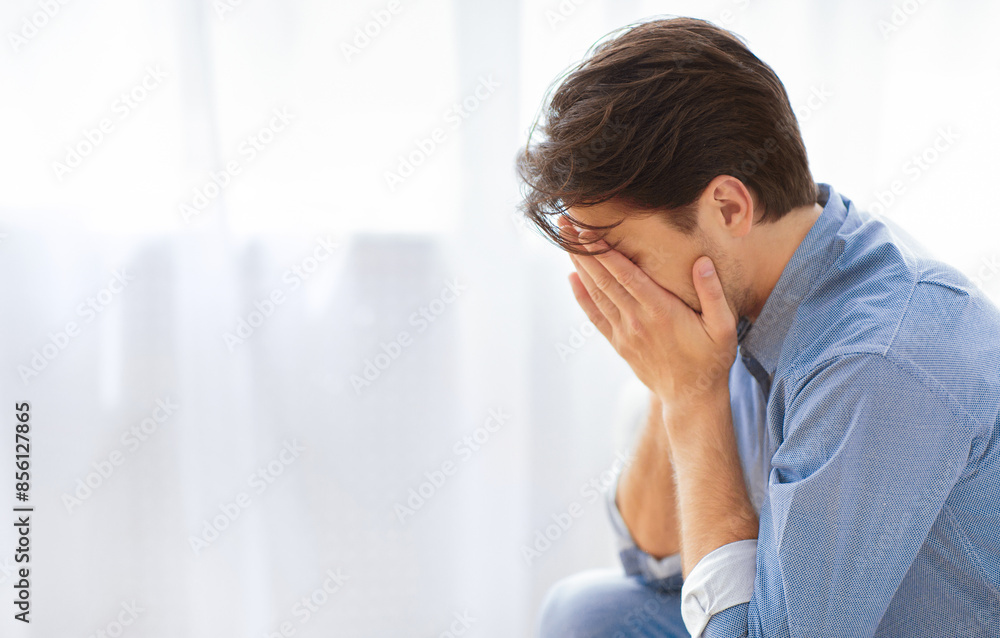 A man sits in front of a window, head bowed and hands covering his face, expressing sadness or despair. A man sits in front of a window, head bowed and hands covering his face, expressing sadness or despair.