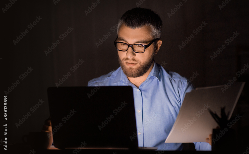 A man wearing glasses and a blue shirt is working late at night in a dimly lit office. He is concentrating on both his laptop and a document he is holding in his hand. A man wearing glasses and a blue shirt is working late at night in a dimly lit office. He is concentrating on both his laptop and a document he is holding in his hand.