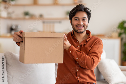 A young man, wearing a red shirt, is holding a cardboard box in front of him in a living room. He is smiling and looking directly at the camera. There is a white couch behind him. photo