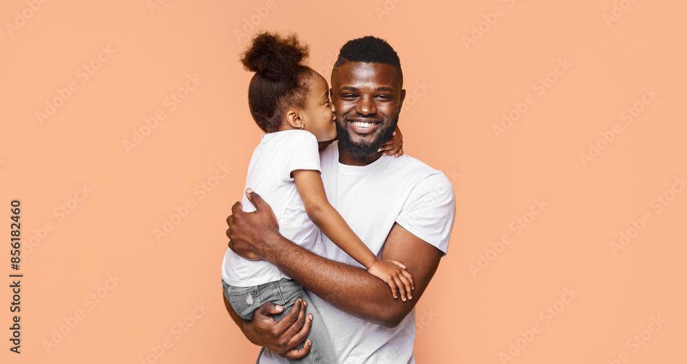 Obraz premium A happy Black father is holding his little daughter and smiling at the camera. They are standing against a peach-colored background