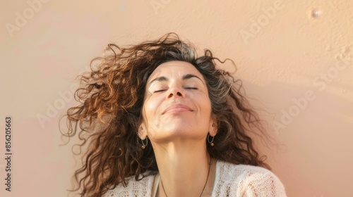 A woman with curly hair closed eyes and a serene expression leaning against a wall with her head tilted back. photo