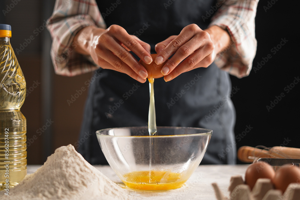 Close-up of female hands separating an egg yolk from the white over a clear bowl in the kitchen. The person is wearing a plaid shirt and a dark apron, Close-up of female hands separating an egg yolk from the white over a clear bowl in the kitchen. The person is wearing a plaid shirt and a dark apron,