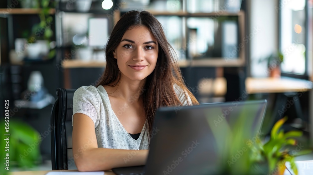 Fototapeta premium Portrait of a young woman in a modern office, seated at a desk with an empty laptop screen, engaged in digital marketing planning, representing her professional management and communication abilities