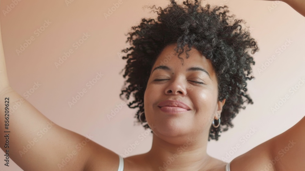 A woman with curly hair smiling and with her eyes closed lifting her arms in a joyful or celebratory gesture against a soft-focus pink background.