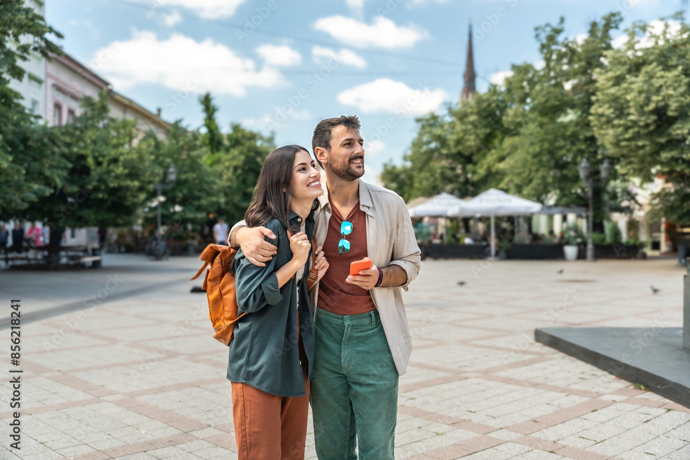 Fototapeta premium Young happy hipster backpacker travelers couple, visiting new country, summer travel destination, enjoying their vacation, walking down the city streets, looking historical buildings and architecture.