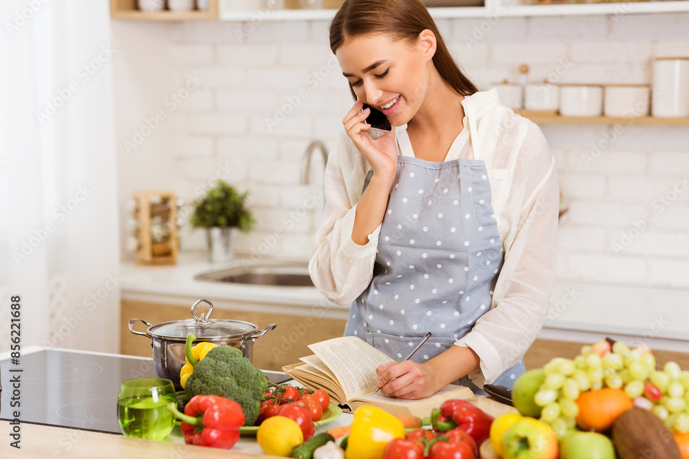 A woman in an apron talks on her phone while preparing a meal in her kitchen. She is smiling and seems happy to be cooking. A woman in an apron talks on her phone while preparing a meal in her kitchen. She is smiling and seems happy to be cooking.