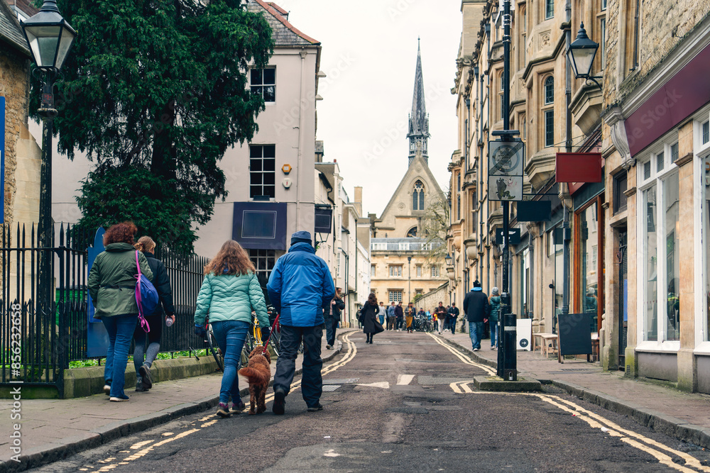 Naklejka premium People walking with a dog on a charming historic street lined with buildings and trees. Serene urban scene capturing everyday life and community, themes on lifestyle travel and urban exploration