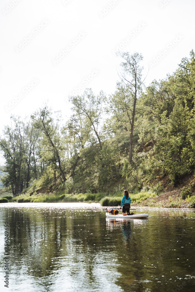 In a peaceful setting, someone is enjoying paddle boarding on a serene river surrounded by the beauty of nature, with water, sky, and trees harmoniously blending in the landscape In a peaceful setting, someone is enjoying paddle boarding on a serene river surrounded by the beauty of nature, with water, sky, and trees harmoniously blending in the landscape