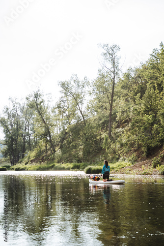 In a peaceful setting, someone is enjoying paddle boarding on a serene river surrounded by the beauty of nature, with water, sky, and trees harmoniously blending in the landscape © Aleksey