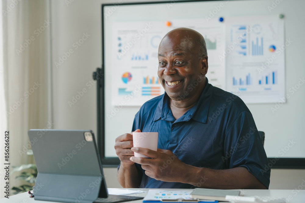 Obraz premium Smiling man drinking coffee and working on laptop at home office. Charts and graphs displayed on board in background. Remote work concept.
