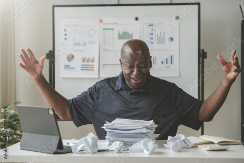 Stressed man surrounded by crumpled papers and documents, expressing frustration in a cluttered office environment. photo