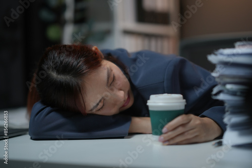 Woman asleep at desk with coffee cup in hand, surrounded by papers. Concept of exhaustion, overwork, and need for rest. photo