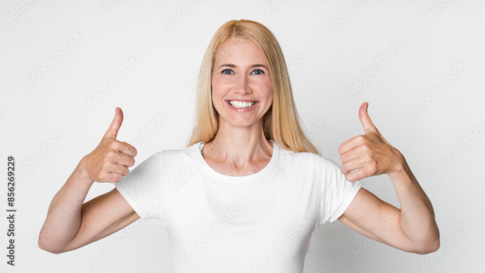 A photo of a smiling blonde woman wearing a white t-shirt against a plain white background. She is giving a thumbs up with both hands. Her smile is genuine and inviting A photo of a smiling blonde woman wearing a white t-shirt against a plain white background. She is giving a thumbs up with both hands. Her smile is genuine and inviting