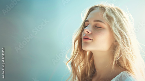 A young woman with long blonde hair enjoys a tranquil moment with her eyes closed against a soft, light blue background, basking in the gentle sunrise. © VK Studio