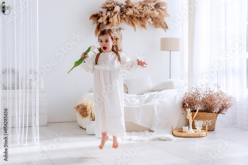 Little girl in white dress, nightgown jumping with back, faceless, on shoulders is hand-made backpack made of raffia with tulips. Light airy, spring interior in rustic style. Pampas grass on the wall