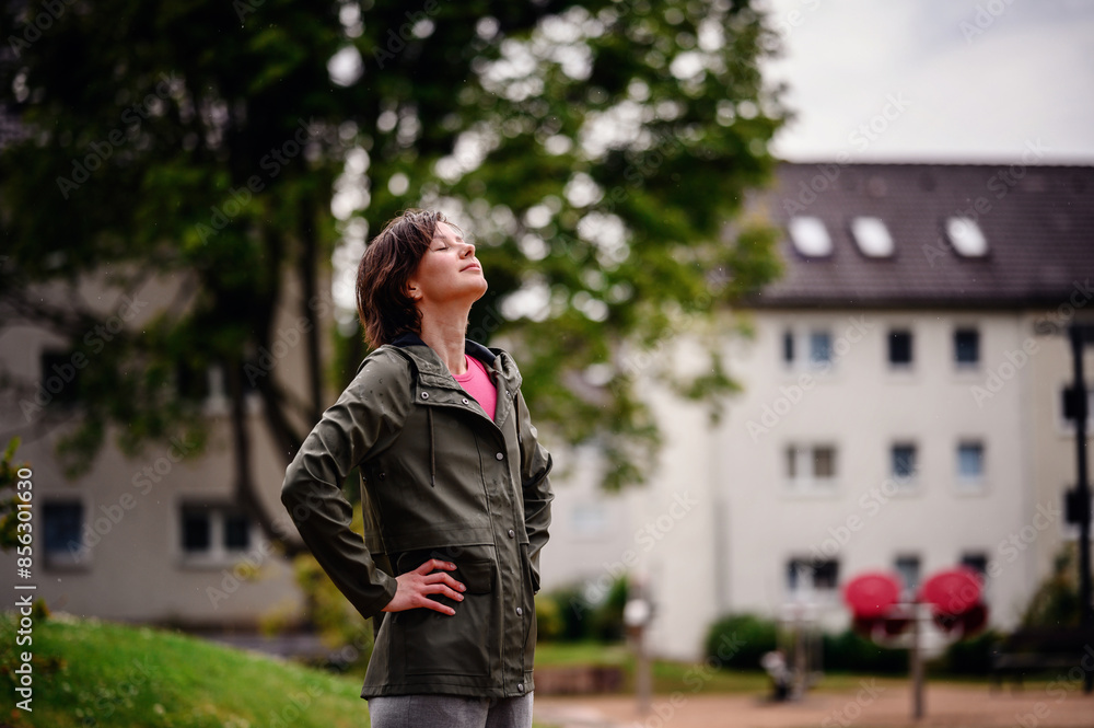 Fototapeta premium Young woman in a green jacket, standing outdoors on a rainy day with a thoughtful expression. The background features a residential area with greenery.