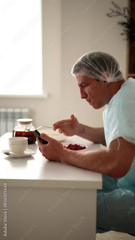 Vertical side view of handsome young man in medical cap with baldness problem resting, eating, and using phone during break in lengthy medical hair transplant procedure. Concept of beauty, healthcare. Vertical side view of handsome young man in medical cap with baldness problem resting, eating, and using phone during break in lengthy medical hair transplant procedure. Concept of beauty, healthcare.