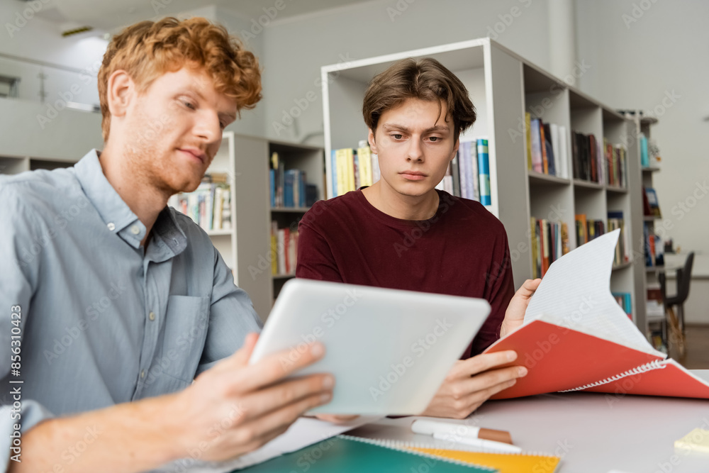 Two students engage with a tablet while seated at a table in a library. Two students engage with a tablet while seated at a table in a library.