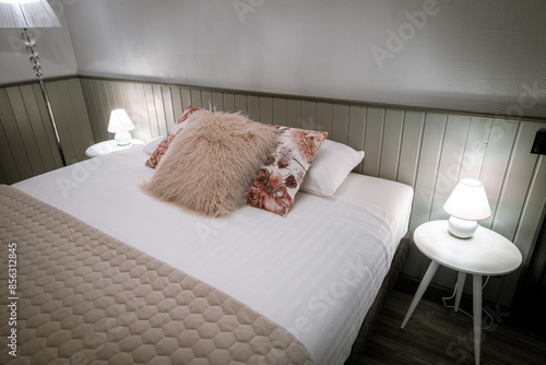 Blome, Latvia - September 11, 2023 - A cozy bedroom with a neatly made bed, floral and furry pillows, bedside tables with lamps, and a soft beige quilted blanket. photo